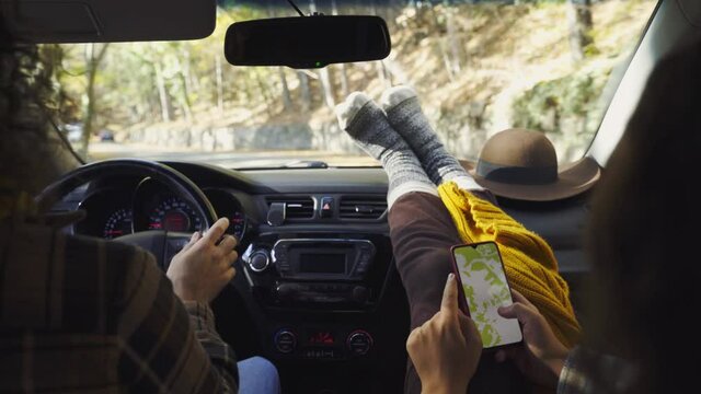 Man drives car along empty rural road across autumn forest while girl friend reads map on phone putting feet on panel closeup from inside salon
