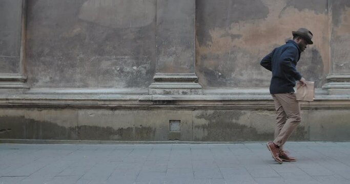 Stylish afro american man in hat dancing along city street. Joyful bearded man in fashionable clothes doing moonwalk while carrying paper coffee cup and craft bag. Outdoors