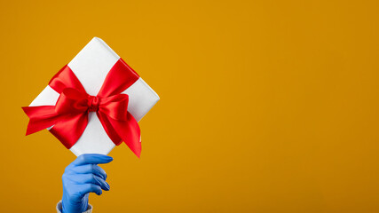 Present surprise. Covid-19 holiday. Valentine day greeting. Hand in protective gloves holding gift box taped red ribbon isolated on yellow copy space. Sending love. Stay safe.