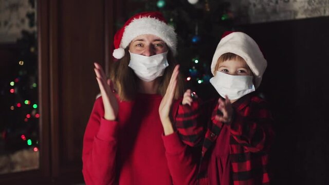 coronavirus, covid-19 in the new year. a little daughter with mother in a red Santa Hat and medical mask is dancing under the Christmas tree at home.