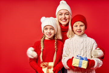 cheerful happy woman with daughter and son holding gifts for New Year 2021, caucasian blonde mother hugs kids isolated over red background
