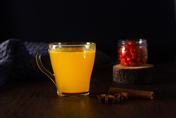 Hot tea or punch with sea buckthorn berries and spices in a glass, jar with sea buckthorn berries on the dark wooden table. Healthy drink, cozy winter night.