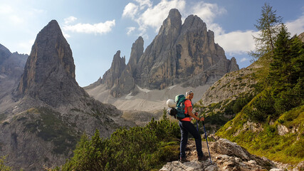 Naklejka premium A woman with big hiking backpack hiking in high Italian Dolomites. There are many sharp peaks behind her. She is enjoying the hike, smiling. There are a few trees around. Sunny day. Outdoor exercising