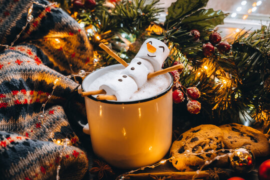 Christmas food white cup with hot chocolate snowman marshmallows. homemade cookie, cinnamon sticks fir xmas tree branches with warm garland lights. New year celebration. Festive dessert breakfast idea