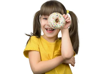 Poster Foto van een aantrekkelijk klein meisje met lange rechte paardenstaarten houdt een geglazuurde witte donut in een hand. Gelukkig meisje dat door de donut kijkt, draagt een casual geel shirt op geïsoleerde achtergrond © Albert Ziganshin