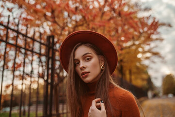 Outdoor close up autumn portrait of young elegant woman wearing stylish orange hat, trendy round earrings, posing in street. Copy, empty space for text