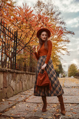 Elegant fashionable woman wearing stylish orange hat, turtleneck, checkered midi skirt, high boots holding suede bag with fringe, posing in street.  Outdoor autumn fashion full-length portrait