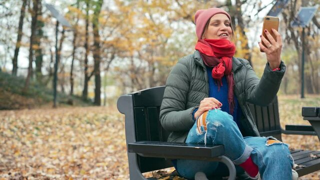 A young woman in a jacket and a hat talking on a video call on her smartphone while sitting on the bench in the autumn park.