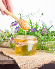 Glass jar of honey and a bouquet of wildflowers on a wooden table on a white background. Honey dripping from the honey dipper into a glass jar of honey. Close up. Copy space. 