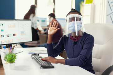 Pov of african businesswoman saying hello while having online meeting wearing face mask as safety precaution against covid19. Woman speaking with team during online conference while colleagues working