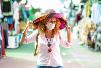 Attractive blonde curly mature woman at flea market wearing a large multi colored hat handicrafted -  wearing surgical mask due to coronavirus infection