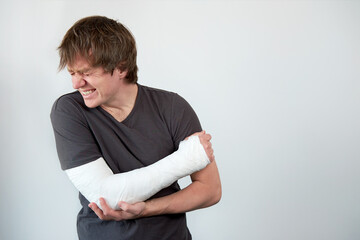 Young unhappy caucasian man with plaster cast on his hand suffering from pain. Shot on a white wall...