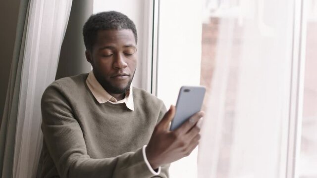Lockdown of young African man wearing casual clothes sitting on windowsill with mobile phone in his hand and talking to friend by video connection