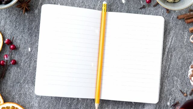 christmas and seasonal drinks concept - snow falling over notebook or diary with pencil, hot mulled wine, orange slices, raisins with cashew nuts and aromatic spices on grey background