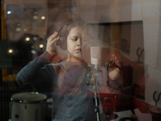 Boy in headphone is singing or talking into microphone with pop filter in voice recording studio. Young singer recording his voice for phonogram performance. View through the window in studio.