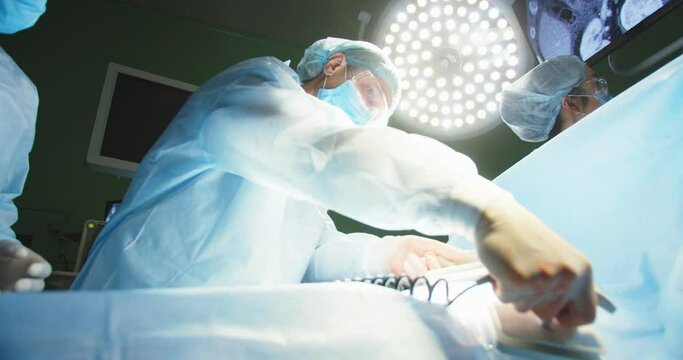 Close up of team of professional doctors in operating room help on intensive care patient, making cpr and use defibrillator electrical device to shock heart under bright lamp. Surgeons and nurses