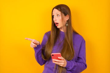 Stunned Young beautiful Caucasian woman wearing purple sweater against yellow wall points sideways right copy space, recommends product, sees astonishing thing
