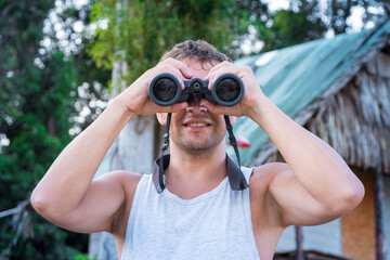 Front view of a satisfied young man in a white T-shirt looks through binoculars against the background of a village hut