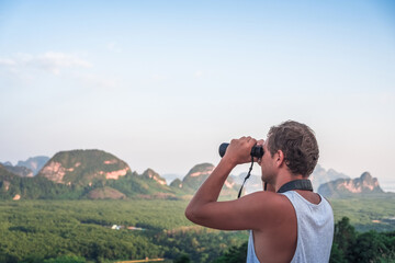 A young man in a white T-shirt looks from the top into the distance through binoculars against the backdrop of wildlife, sky, mountains and sea