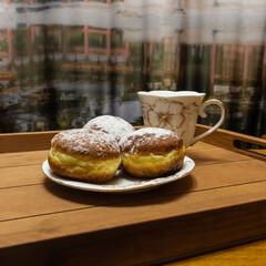There is a saucer of donuts and a cup on a beige wooden tray. The pastries are sprinkled with powdered sugar. The dishes are white with a golden floral pattern. Abstract background.