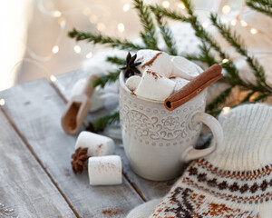 Obraz na płótnie Canvas Winter hot drink. Cozy home composition with white mug with chocolate and marshmallow, cinnamon. Knitted mittens, christmas lights, wooden background. Festive holiday atmosphere, family spirit. Close