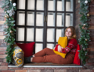 A blonde girl in a red sweater sits on the windowsill.