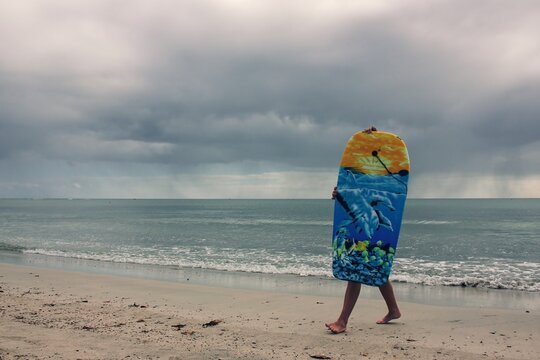Disappointed boy surfer holding surfboard while leaving on beach hiding from ocean wind to wait rain thunderstorm ending and continue surfing. Kids swimming with body board.Active family lifestyle