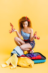 curly woman puffing cheeks while holding shoes and looking at suitcase with clothing on yellow
