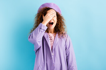 scared and curly woman in beret covering face with hands and screaming on blue