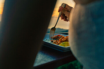 Hands of a young woman sitting at wooden table and enjoying food in a restaurant on the top place at sunset.