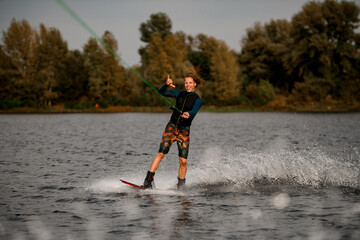cheerful young man holding cable and rides wakeboard showing hand gesture.