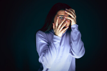 Hiding. Portrait of young crazy scared and shocked caucasian woman isolated on dark background. Copyspace for ad. Bright facial expression, human emotions concept. Looking horror on TV, cinema.
