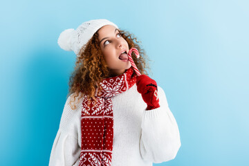 curly young woman in red mittens licking candy cane on blue