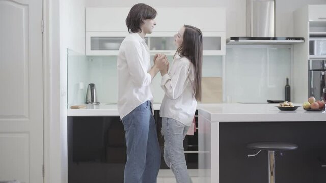 Wide shot of smiling boyfriend and girlfriend rubbing noses in kitchen at home. Side view of happy loving young man and woman enjoying weekend morning indoors. Bonding and love concept.