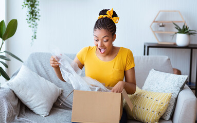 Excited black woman unpacking box after online shopping - obrazy, fototapety, plakaty