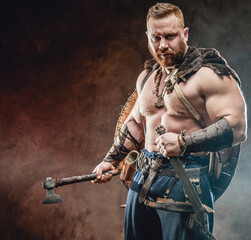 Shirtless and handsome scandinavian warrior with muscular build holding hatchet and knife in smokey background.