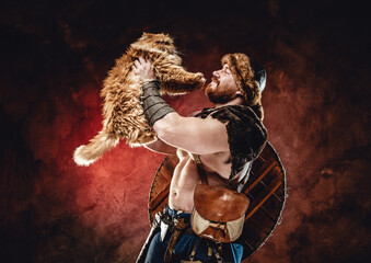 Portrait of joyful and muscular viking warrior with shield on his back he plays with his little friend in dark red background.