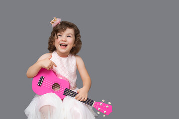 A beautiful and funny little girl playfully playing the pink toy guitar in the studio on a gray...