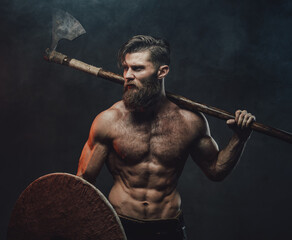Shirtless medieval warlike viking with beard and muscular build posing holding two handed axe in dark smokey background.