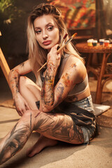 Portrait of creative hipster woman with blond hairs and tattooed leg and arm holding paintbrushes...