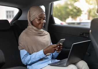 Black islamic businesswoman using smartphone and laptop, going to office by car