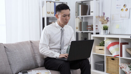 concentrated asian guy worker in suit seated on sofa is preparing financial statement with notebook and document.