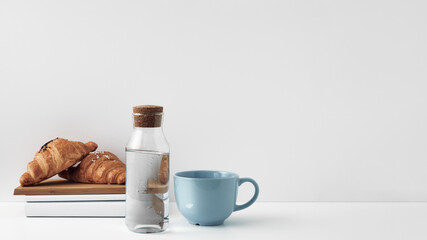 A transparent bottle with a cork stopper, a mug and books with fresh croissants on a white background. Natural and eco-friendly materials in interior decor. Copy space, mock up.