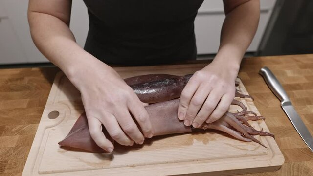 Woman peels squid on a cutting wooden board with a knife in the kitchen in the apartment. Housewife prepares healthy seafood meal. Healthy food for the family. Correct protein