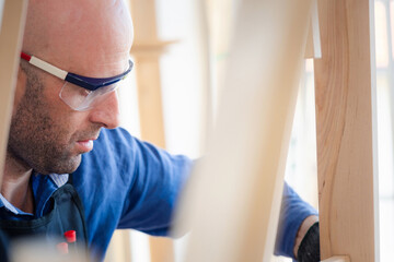 Naklejka na ściany i meble Carpenter worker holding wood plank and check the quality before made furniture in the workstation. A man wearing safety glasses to protect wood chips. Concentrate on work. Handmade, DIY concept