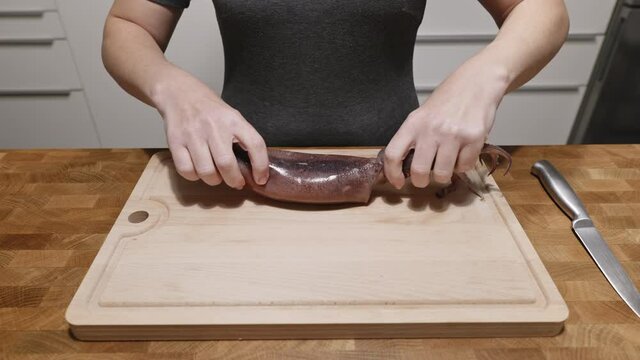 Woman peels squid on a cutting wooden board with a knife in the kitchen in the apartment. Housewife prepares healthy seafood meal. Healthy food for the family. Correct protein