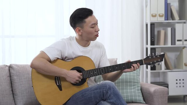 asian man playing the guitar in his free time on living room sofa, and put his foot stamping gently along with the music.