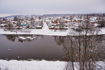 Snow-covered village with colourful wooden houses on the banks of the river, in a cloudy winter day.