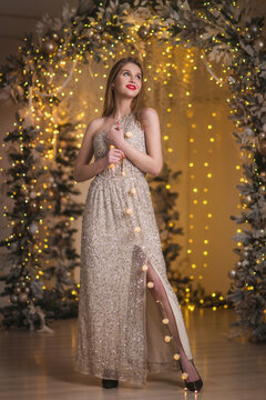 Elegant woman in evening beige fashion dress in golden christmas decorations. Bokeh lights and snowy new year decorated arch. Magic miracle surround full length portrait