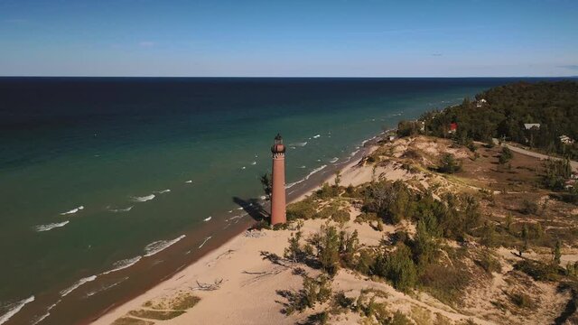 Beautiful aerial view circling around Little Sable Lighthouse along the shoreline of Lake Michigan sitting amongst vegetation and tree covered sand dunes with a clear blue sky above.